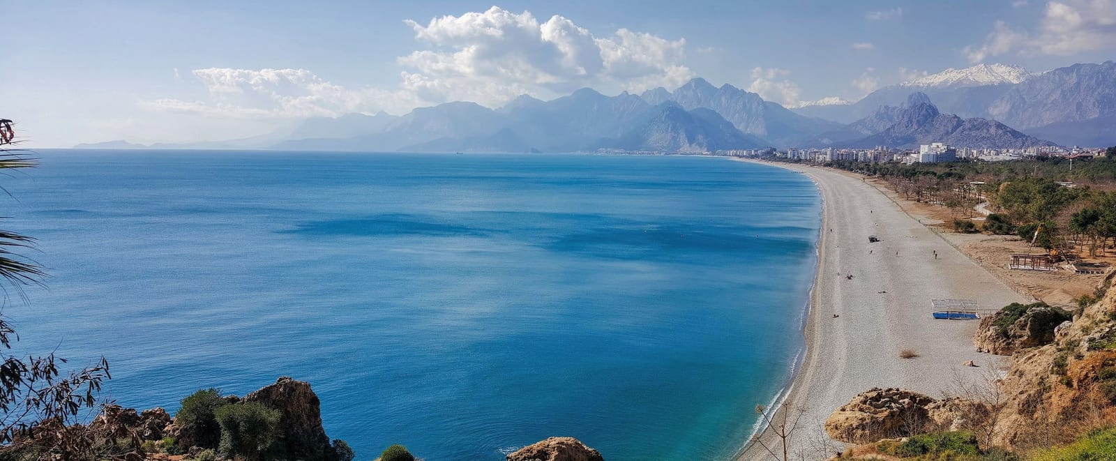Konyaaltı’s pebble beach and blue bay in Antalya, seen from the cliffs, with the city in the distance and the Taurus Mountains behind on a clear morning.